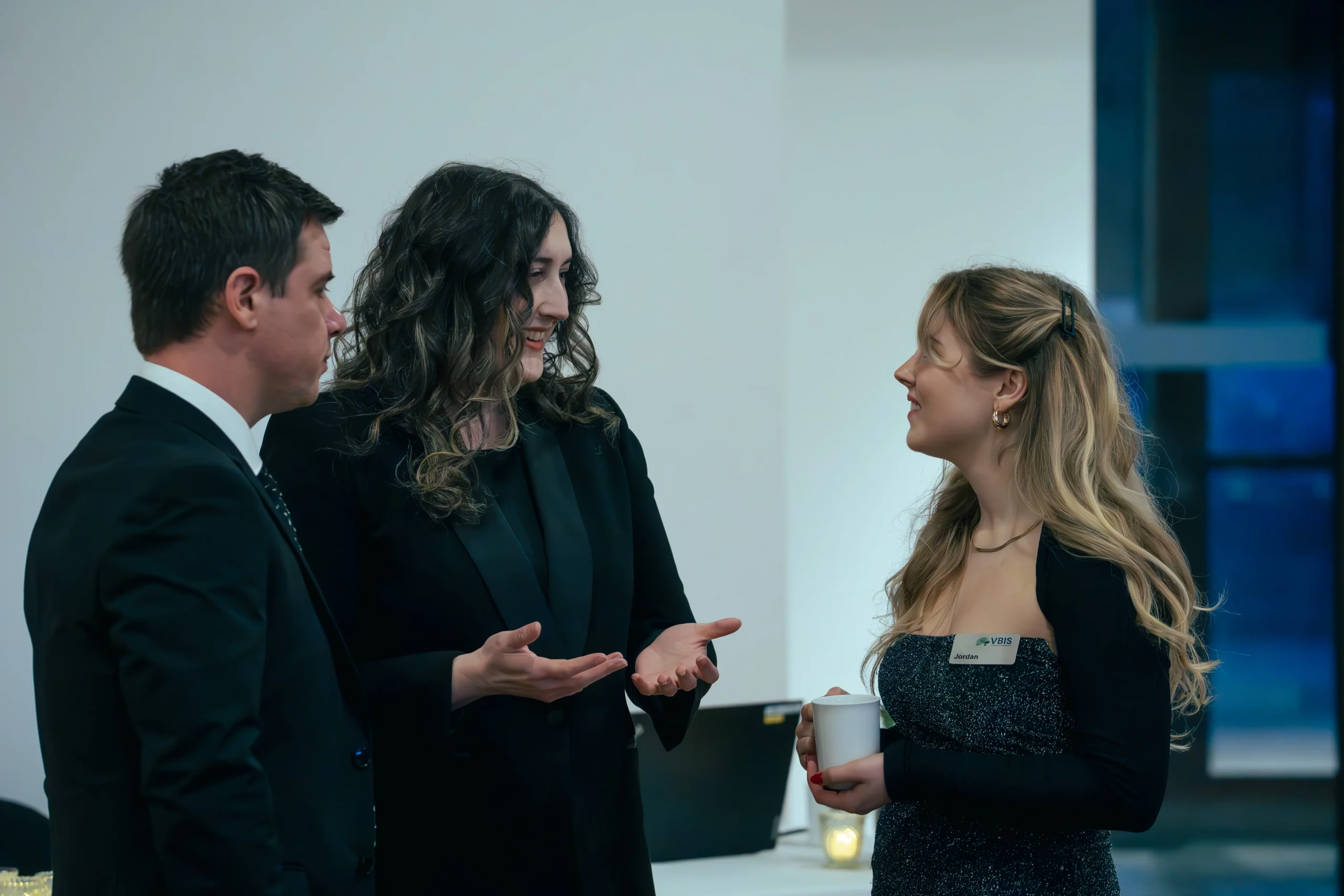 Three people in formal attire engage in a lively conversation at an indoor event. A woman gestures expressively, creating a warm, engaging atmosphere.