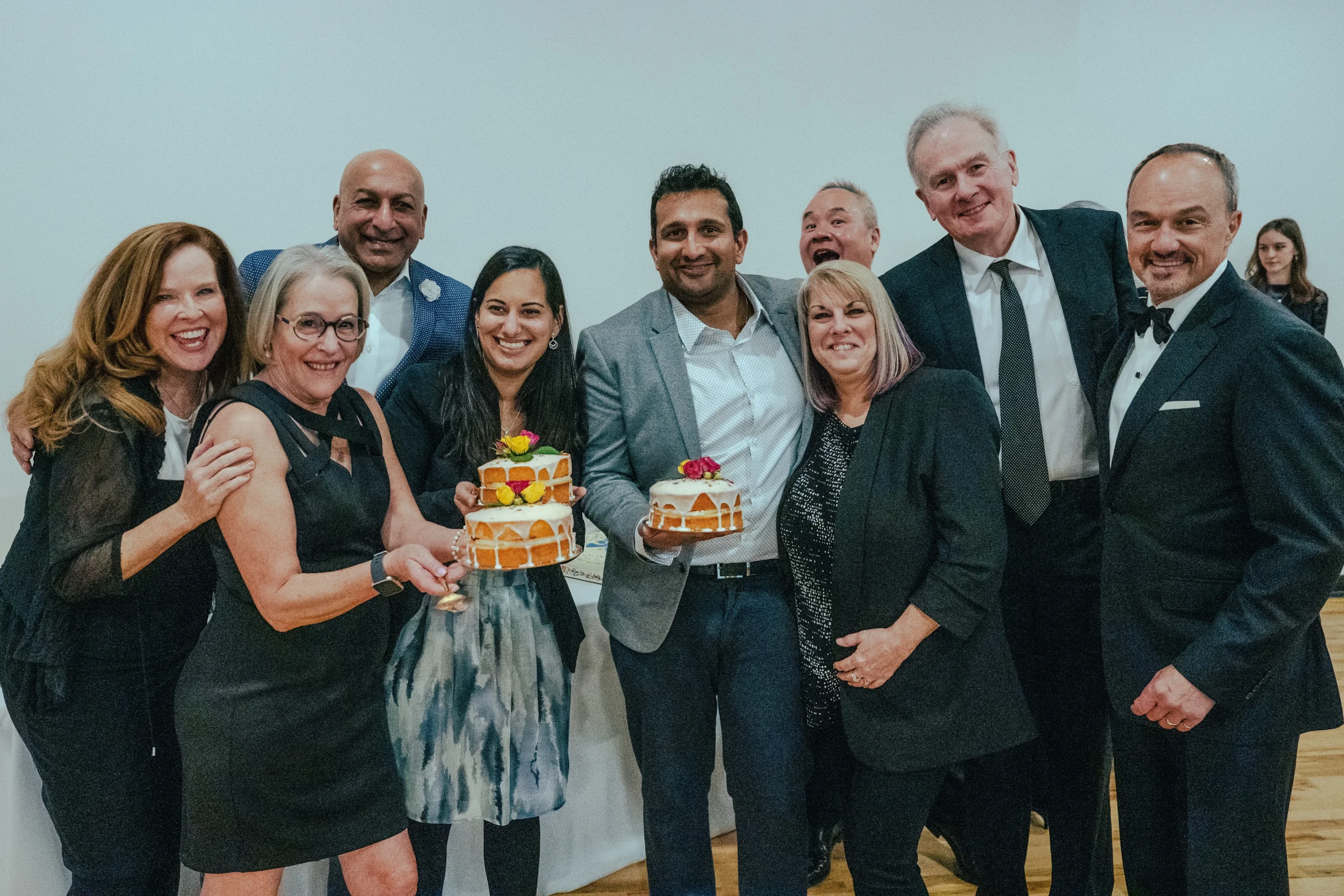 A group of nine people smiling at an indoor event, two holding colorful cakes. The mood is joyful and celebratory, with a casual, friendly atmosphere.