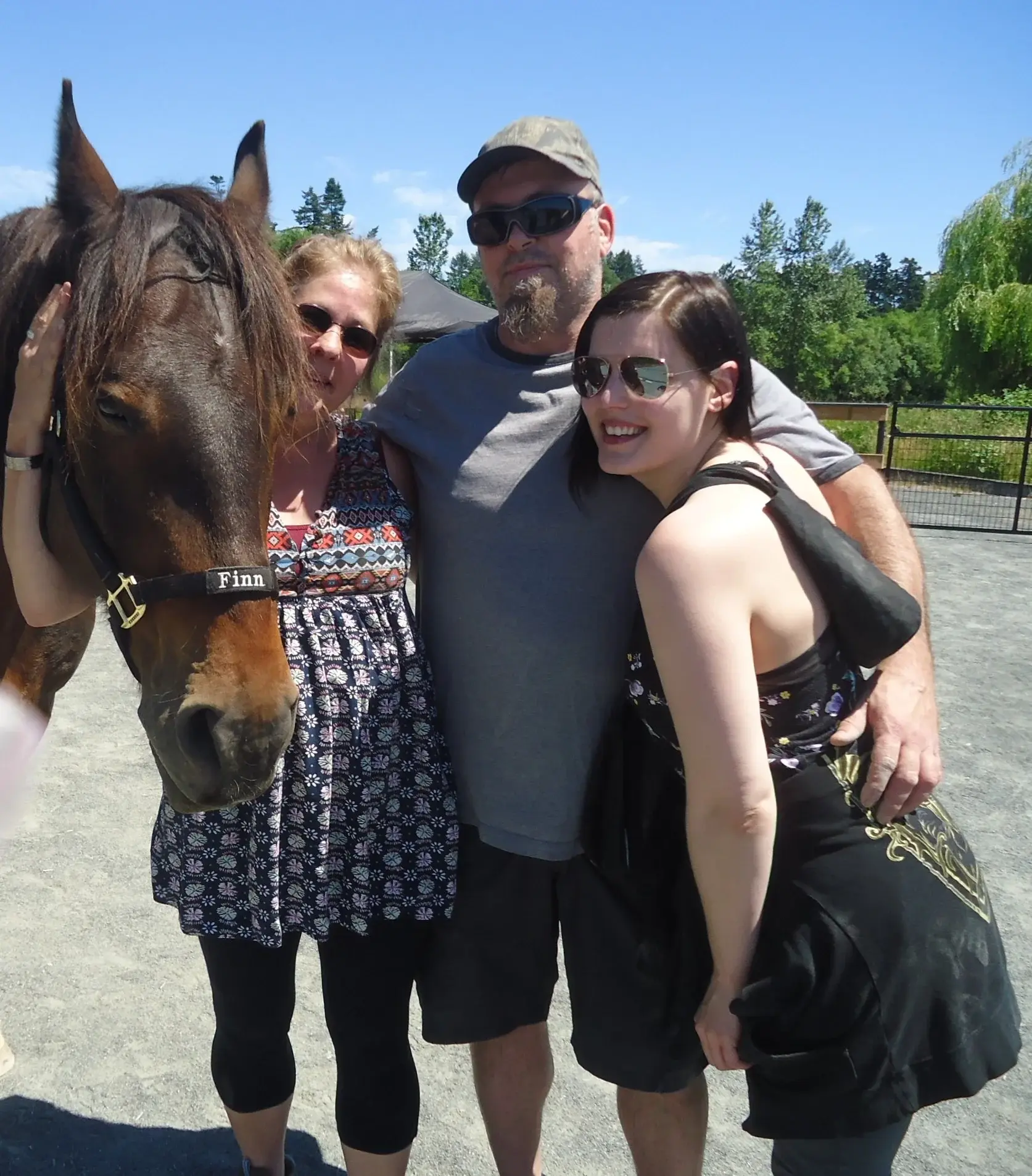 Horse Assisted Learning - Victoria Brain Injury Society A group of three smiling people stand closely together with a brown horse named Finn, outdoors on a sunny day. They appear happy and relaxed.