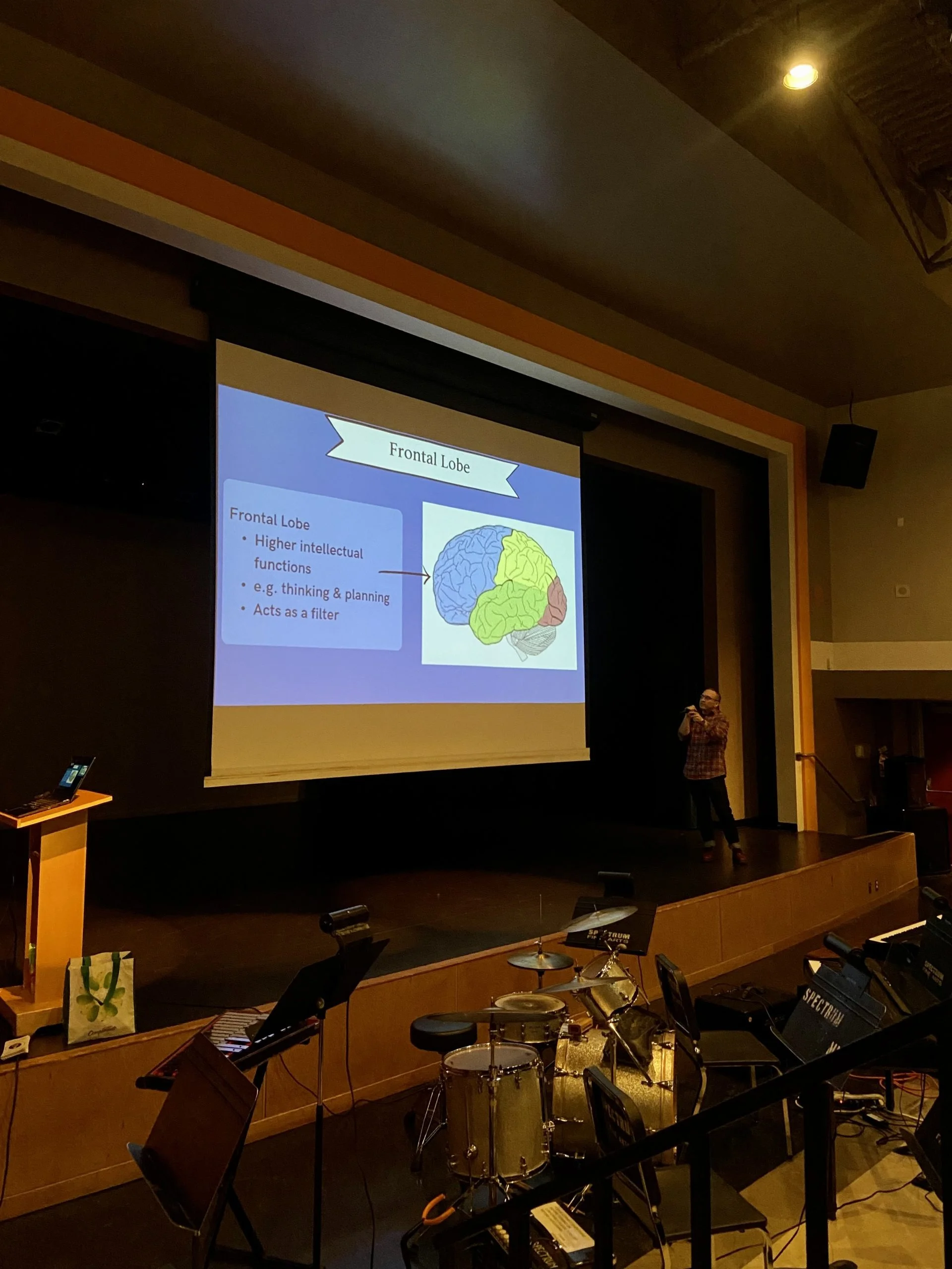 A presenter stands on a dimly lit stage next to a large screen displaying a diagram of a brain labeled "Frontal Lobe," with notes on its functions. Musical instruments are visible in the foreground, suggesting a multipurpose venue.
