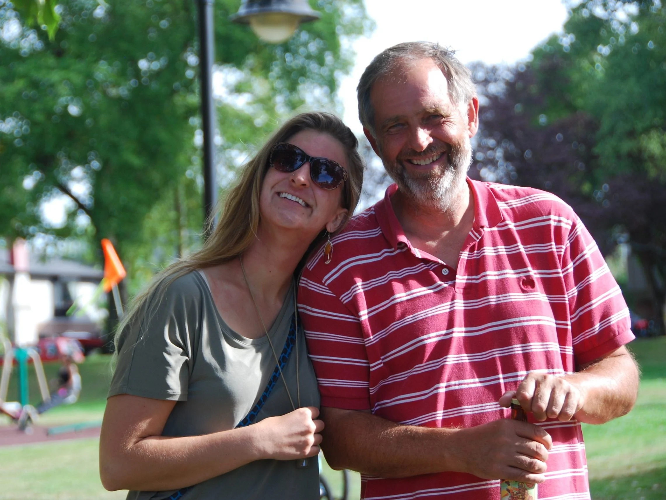 A woman and man smiling outdoors, she leans on his shoulder. She wears sunglasses and a gray shirt, he is in a red striped shirt. Sunny day in a park. 
